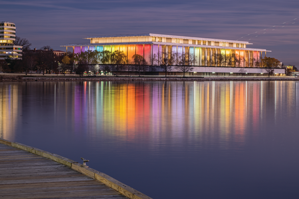 Color HABS photograph at dusk showing the exterior of the Kennedy Center lit with rainbow colored lights. A reflection of the colorful structure can be seen in the Potomac River.