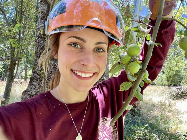 Close up of intern Kennedy Little wearing an orange hard hat, a branch with green leaves hangs to their right