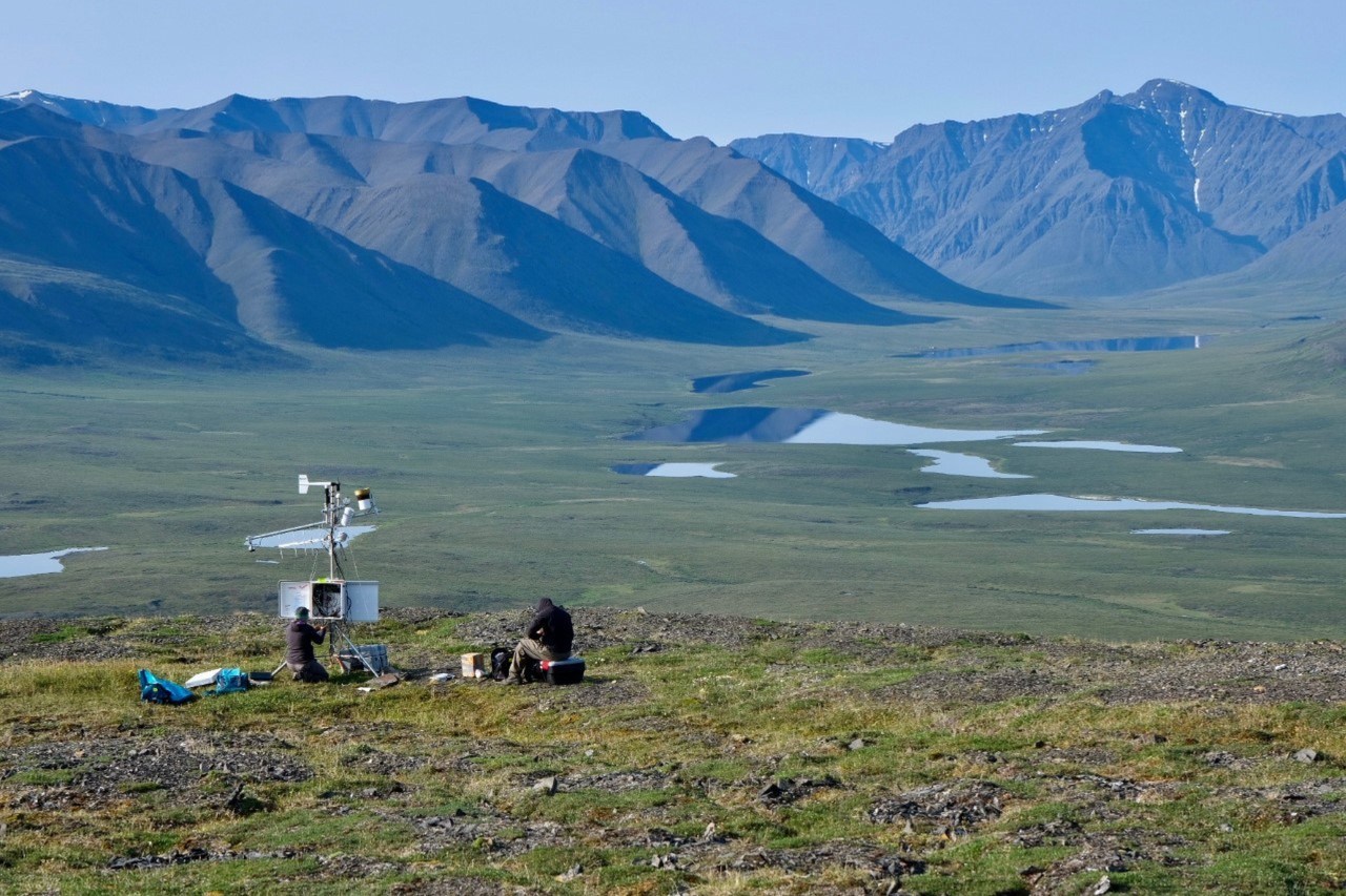 Two people work on maintaining a climate station against the backdrop of a wide tundra valley ringed with mountains.