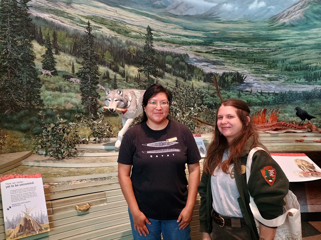 Two people pose for a photo in front of a diorama featuring a wolf carrying a bloodied bone.