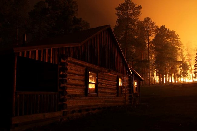 A wildfire blazes behind a historic log cabin at night.
