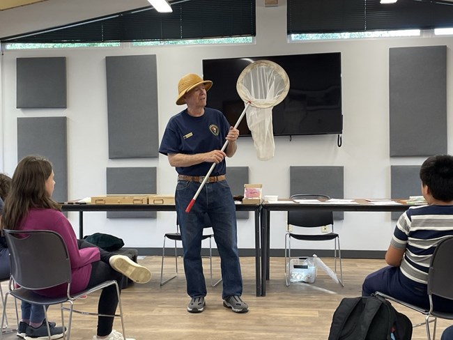 Man in a blue shirt and tan field hat holds a large bug net up to an audience.