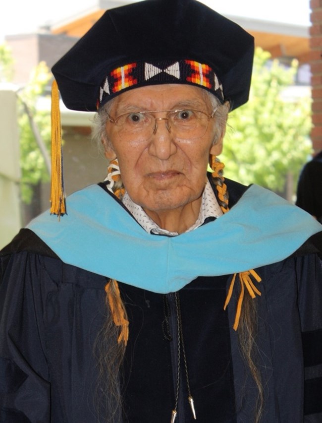Tribal elder wearing black graduation doctoral robe, blue collar, and tam with beaded band.