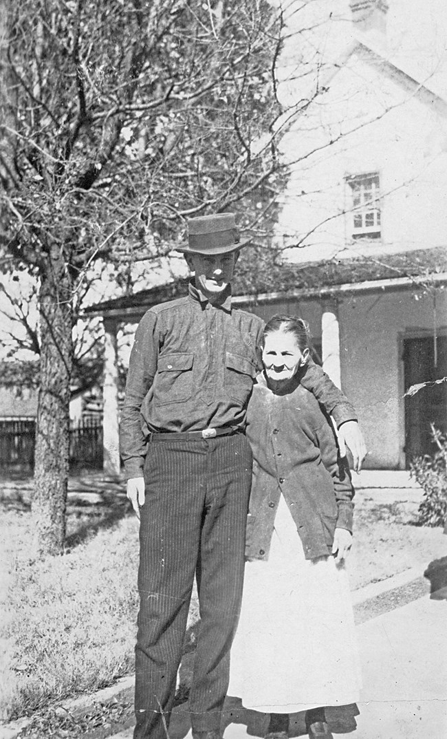 A tall young man stands next to a petite older woman in the front yard of a two-story house.