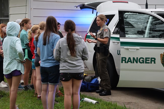 A group of young women listen to a woman wearing a green and gray National Park Service Uniform as she stands in front of a vehicle that says U.S. Park Ranger on the side.