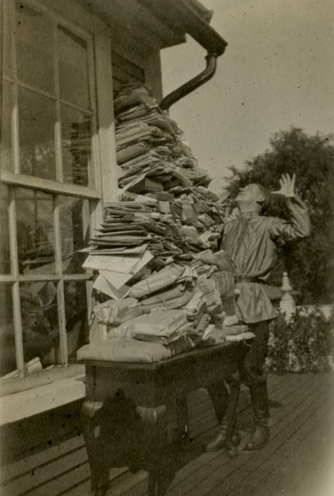 White man wearing a Cossack shirt and tall boots poses with his hands in the air looking up at a pile of papers on a table outside.