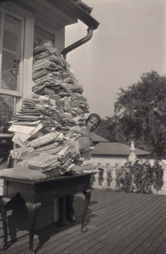 Table piled with documents and packages higher than a person's height, with only the head of a Black man visible peeking around the stack.