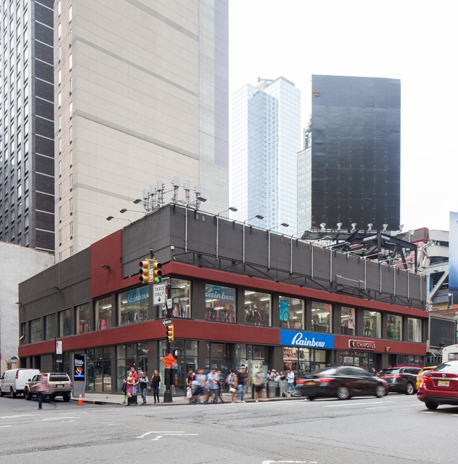A large black multi-story building with maroon banding between floors, sitting on a street corner with signs on its righthand façade reading “Rainbow” and “Chipotle.” Pedestrians are on the sidewalks around the building and cars are driving by.