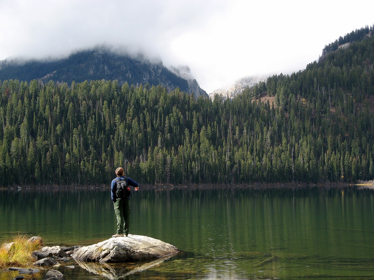 A mountain lake surrounded by forest and steep mountain slopes. A man stands on a rock overlooking the lake.
