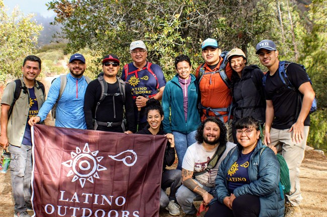 A group of hikers pose for a picture holding a burgundy flag with the Latino Outdoors Logo.