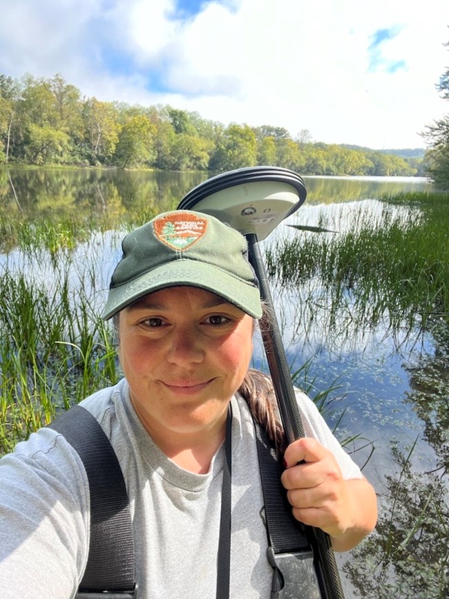 Smiling park ranger holding a GPS surveying device, standing in front of a  lake surrounded by tall green grass