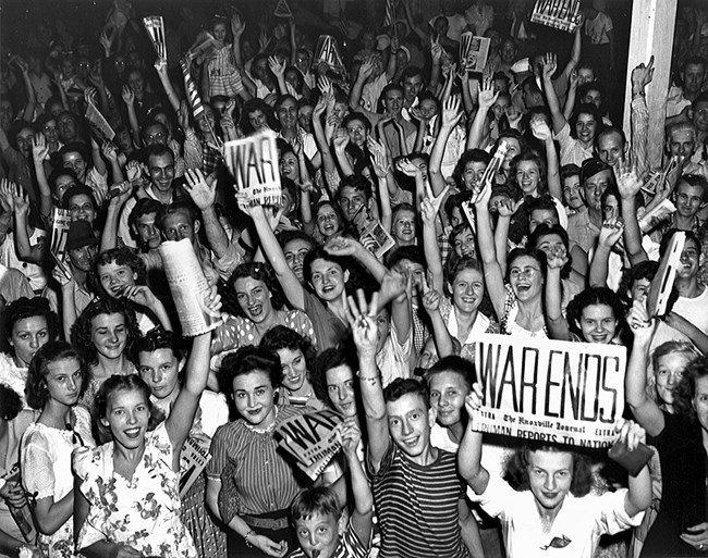 Black and white photo. People – mostly white women – stand shoulder-to-shoulder cheering and waving. Some hold up the front page of The Nashville Journal with the headline “WAR ENDS.”