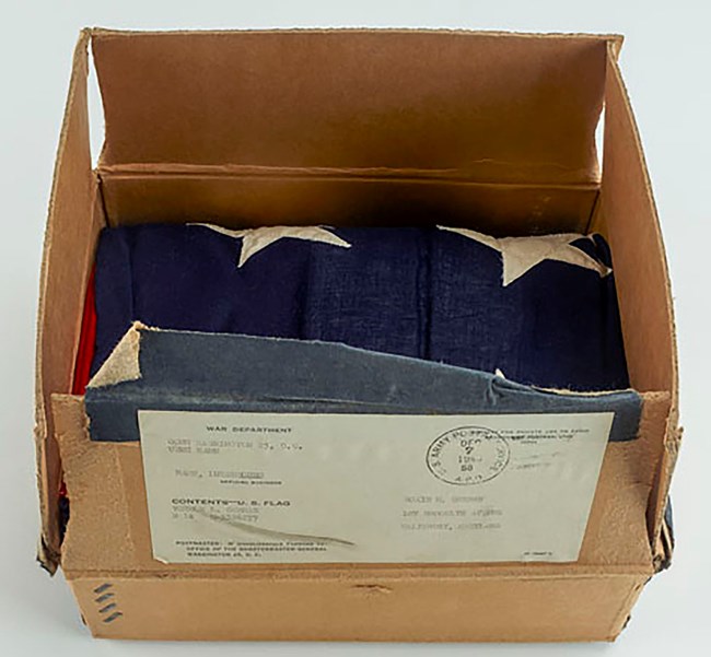Color photo of a folded American flag resting in its original cardboard shipping box.