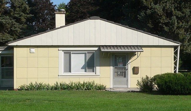 Color photo of a bungalow. The sides of the home are made of butter-yellow steel panels. Trees rise behind, and in front is a lush green lawn.