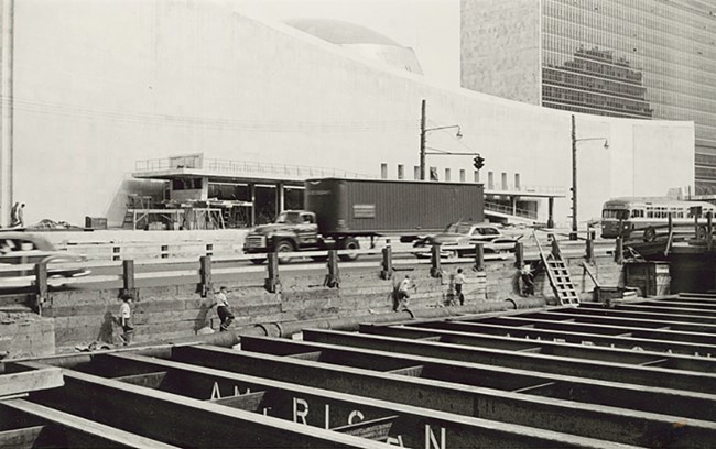Black and white photo. Young boys are dwarfed by the size of the steel girders forming supports for an underground area. Behind them, the curved front of the UN building is mostly completed.