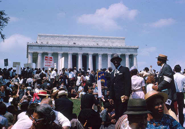 Color photo of African Americans, dressed in suits and dresses, standing and sitting on the lawn of the National Mall. Behind them is the Lincoln Memorial against a blue sky.