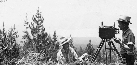 Black and white photo of a man using an large old-fashioned camera on a tripod in an outdoor setting with trees in the background while another man looks on.