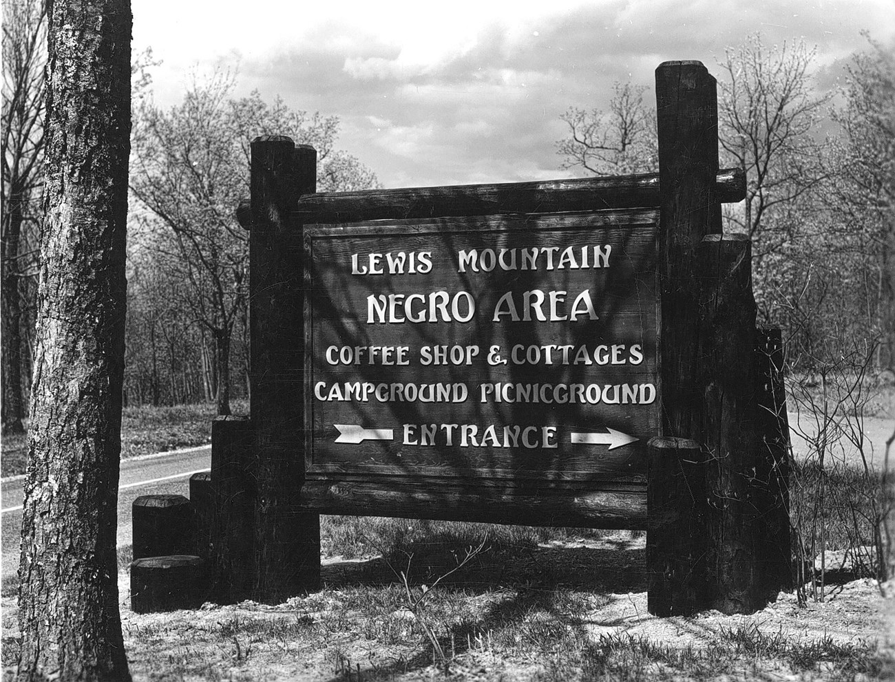 Historic black and white photo of a wooden sign with white lettering reading, "Lewis Mountain Negro Area Coffee Shop & Cottages Campground Picnic Ground." An arrow dissected by the word "Entrance" points to the right.