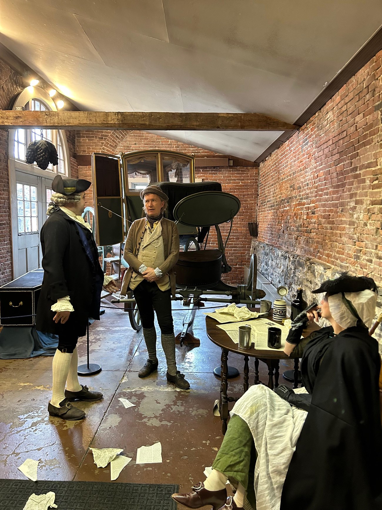 Two men standing in front of a carriage converse as seated woman listens. All individuals wearing colonial-era clothing