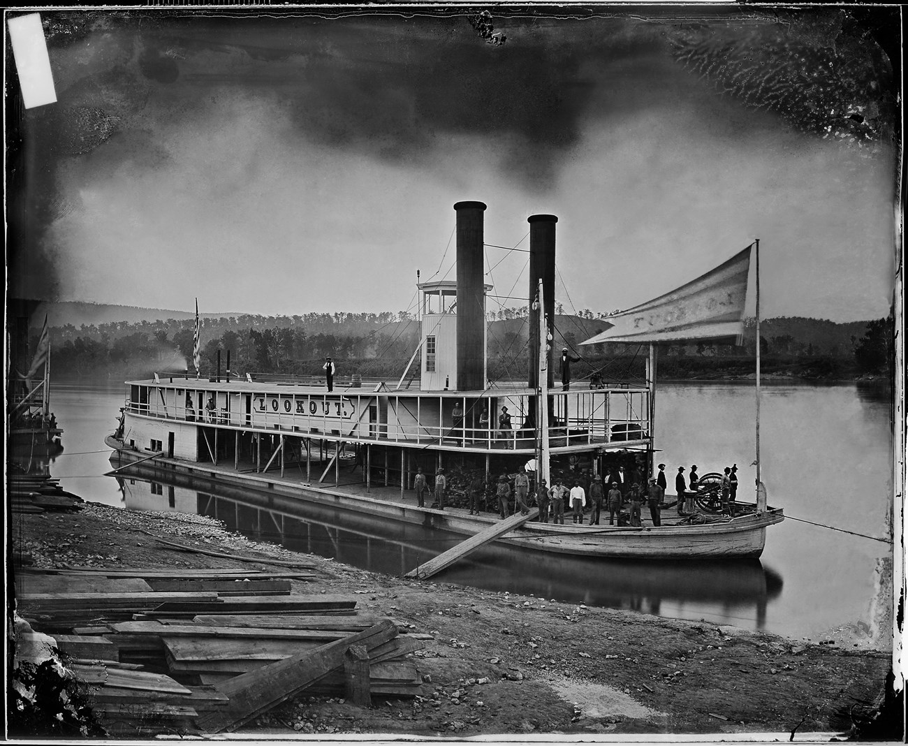 Wooden steamboat with a deck and two chimney stacks sailing down a river. About 20 men are on the steamboat.