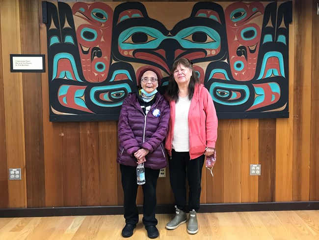 Two women stand in front of a carved panel.