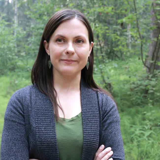 White woman with long brown hair wearing a blue cardigan standing in the woods