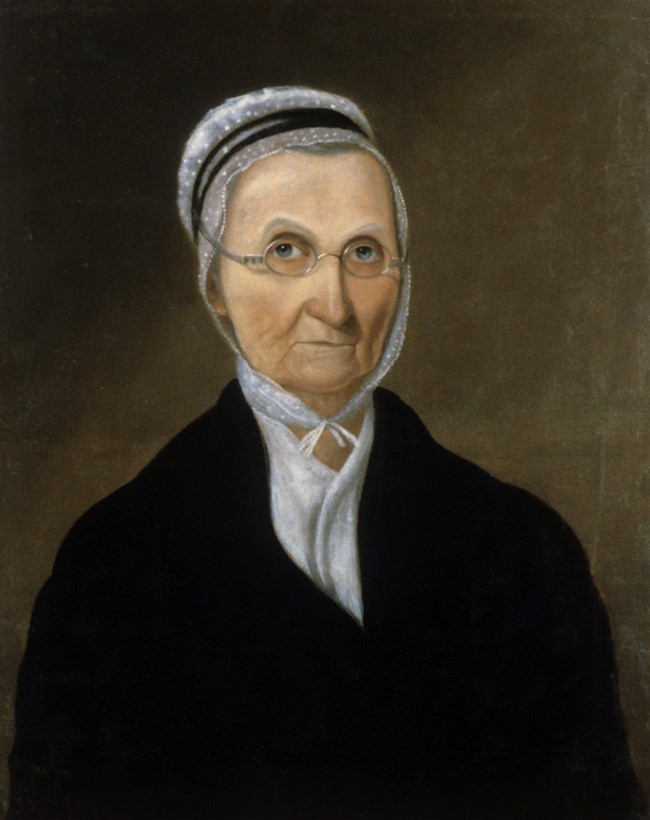 A painting of Lucy Meriwether Lewis Marks. The expressionless woman wears a white bonnet with black trim and small, round glasses. She has a white shirt and black jacket. Her portrait is set against a brown background.