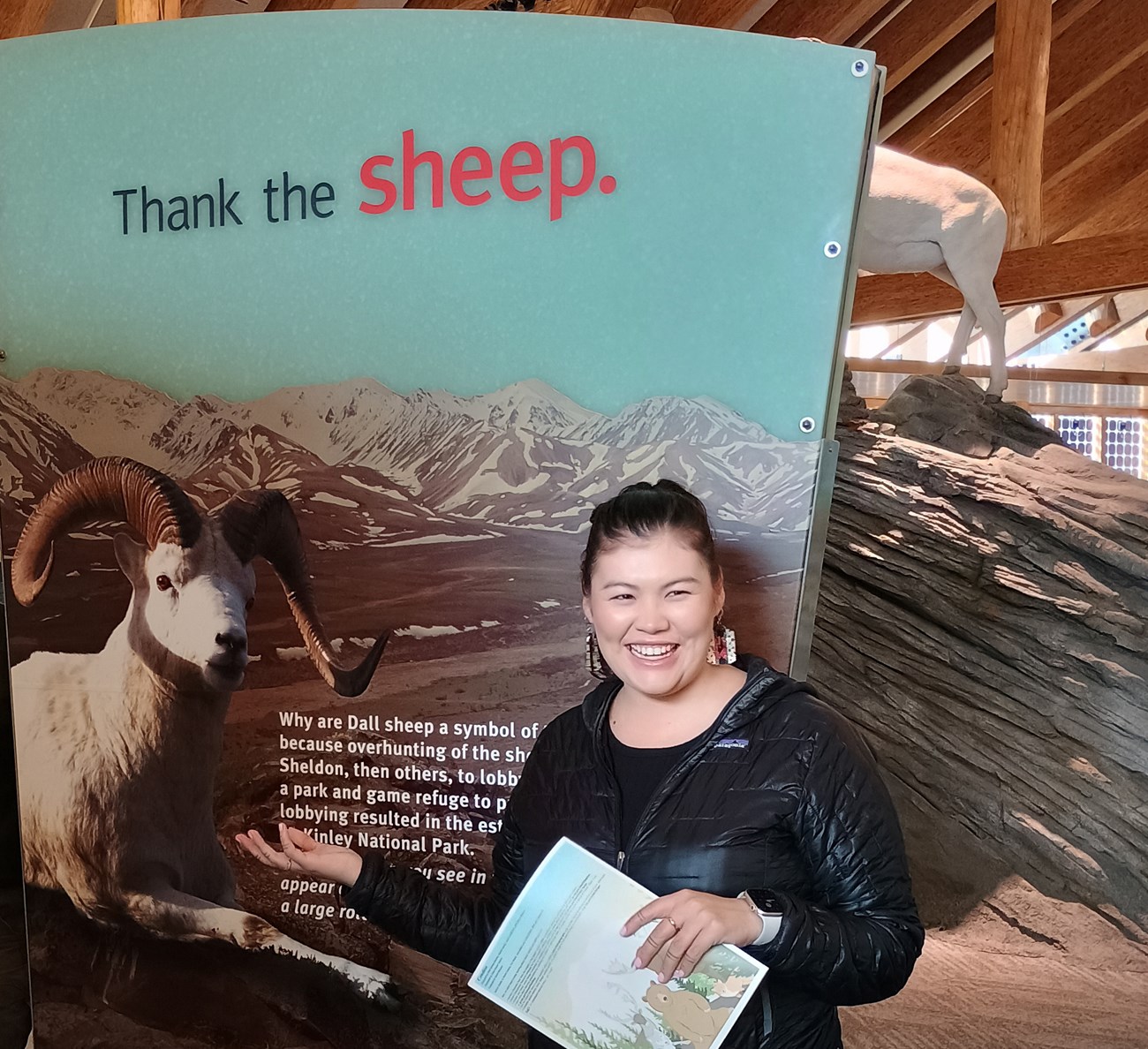 Agnus smiling and gesturing toward a visitor center display featuring a photo of a Dall sheep in the mountains with text that reads "Thank the sheep."