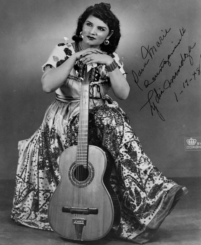 Woman sitting down leaning on a guitar. She wears a dress and the photo is in black and white