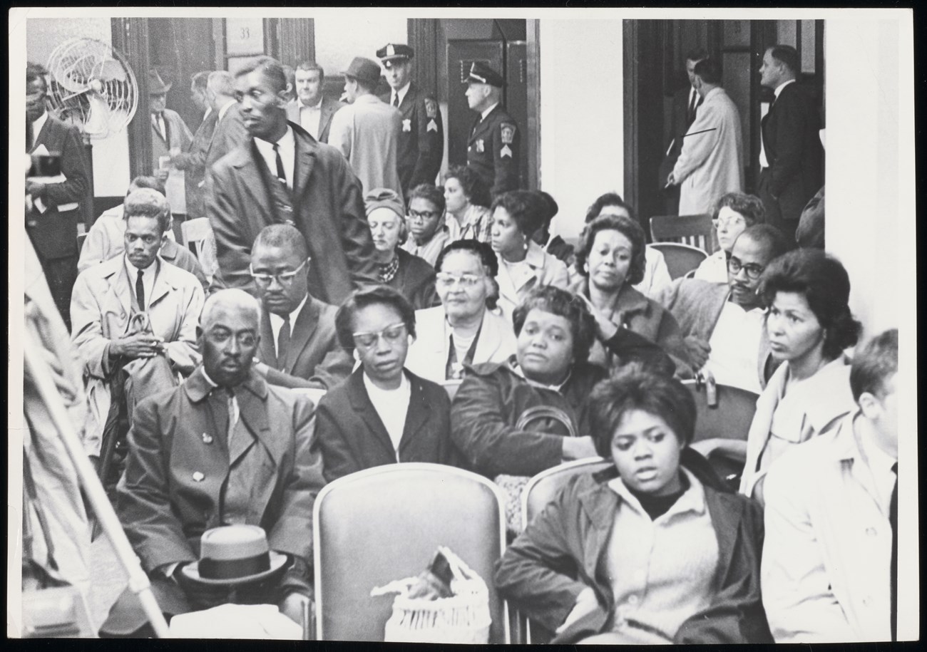 Black and white image of middle aged Black folks sitting in chairs in an auditorium
