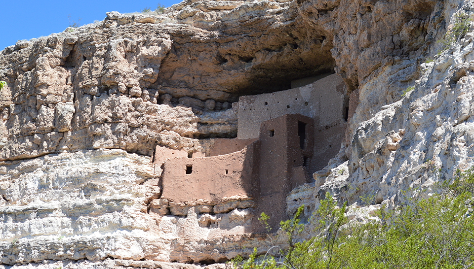 Steep rock cliff faces with a multilevel adobe structure built into a cavernous opening in the cliff.
