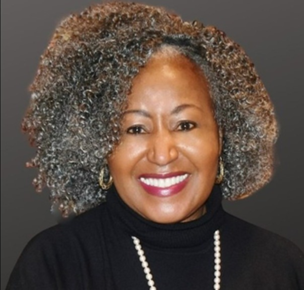 A portrait of a smiling woman with curly hair in a black turtleneck with white pearls.