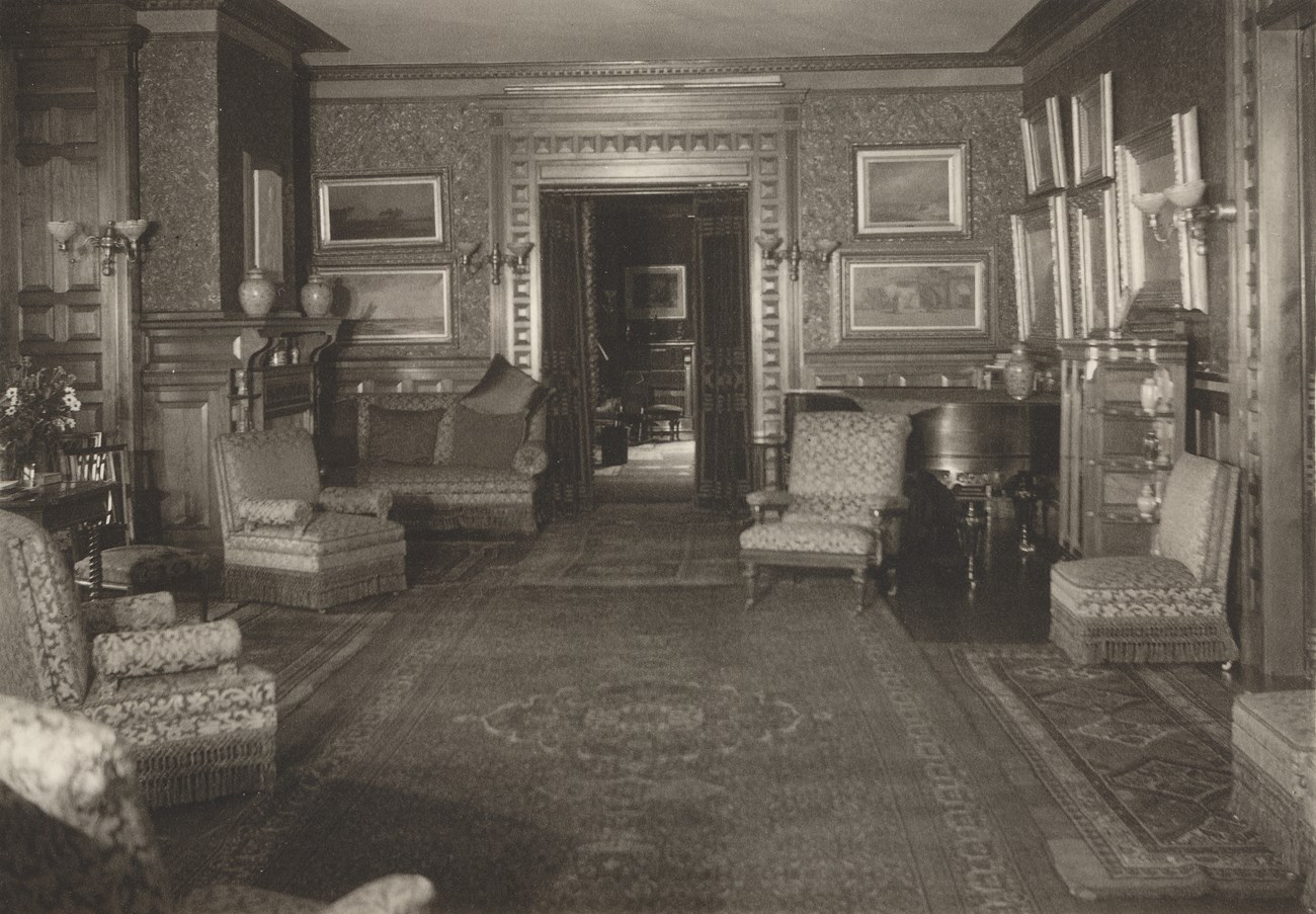 Photograph of Parlor in the Mansion, 1935. The perspective is a eye level looking down the room towards the door into the Hall. On the Left are several chairs, a sofa and the fireplace. The walls are hung with many framed paintings.