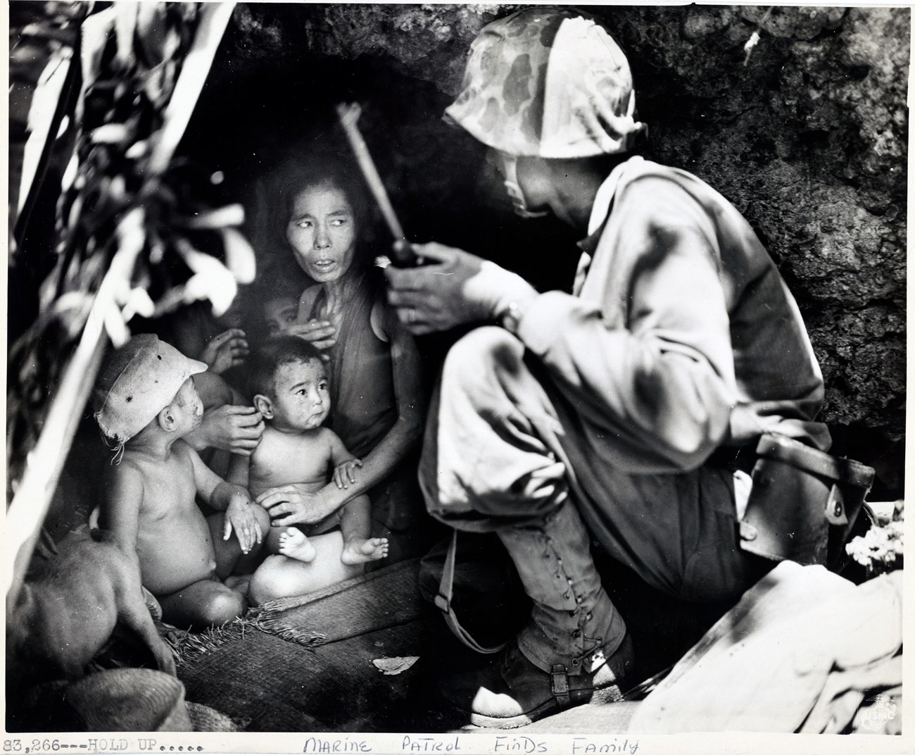 Uniformed Marine kneels at mouth of cave with emaciated woman surrounded by six children
