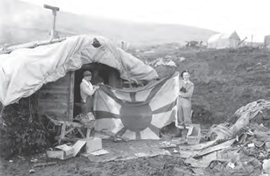 Two man stand outside a tent cabin holding a Japanese flag. The flag has a large circle in the middle with wide lines that radiate outwards towards the edges of the flag. Rolling hills are in the background with another tent cabin on a distant hill.