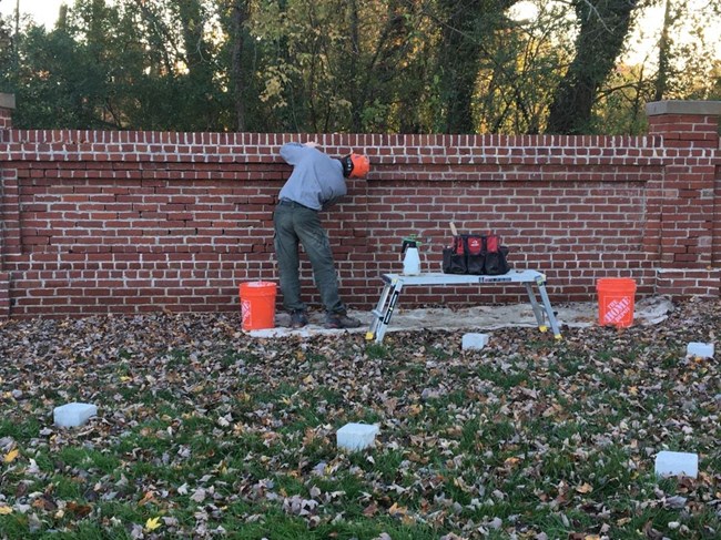 A worker, in an NPS uniform and a hard hat, works on a shoulder height brick wall with a small workbench and tools. Grave markers are evenly spaced in the green grass nearby.