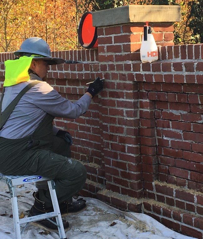 A person in an NPS uniform and hard hat sits on a workbench in front of a brick wall.