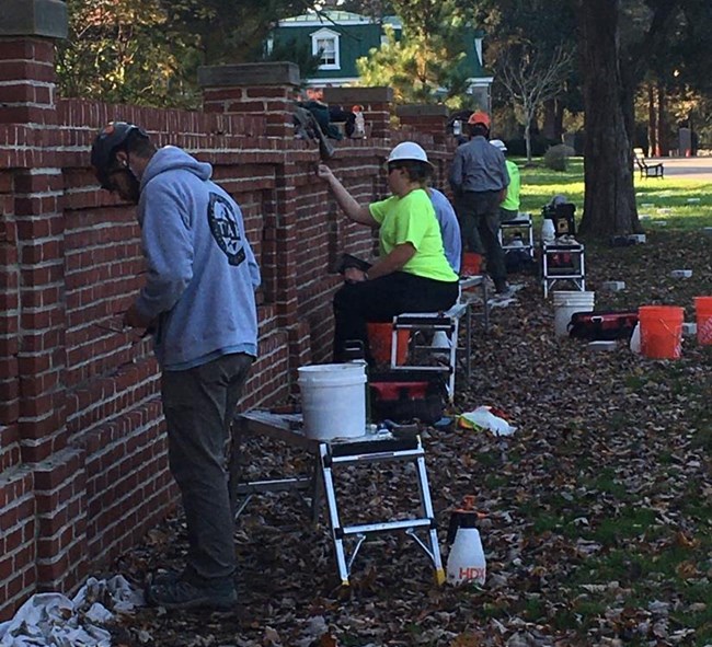 Five people in hard hats work on various sections along a long brick wall.