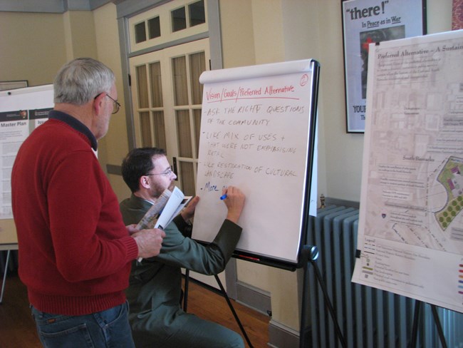 A member of the public stands in front of two posters, one showing a map of the Barracks properties and the other showing comments provided by attendees. A National Park Service ranger kneels in front of the second poster to write additional comments.