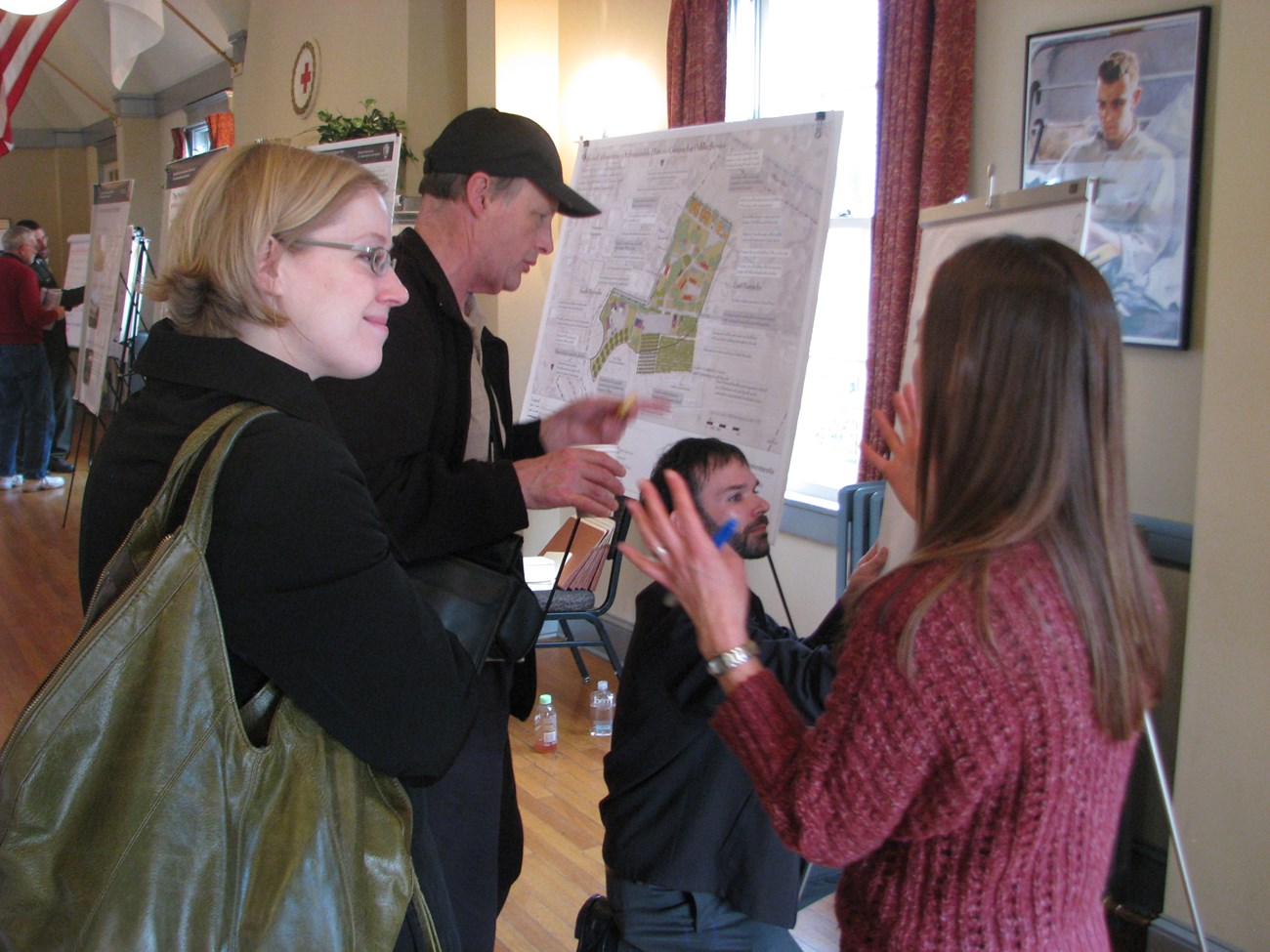 Two National Park Service employees talk with two members of the public. One kneels in front of a comment board, recording comments from the public. In the background is a poster showing a map of East Vancouver Barracks.