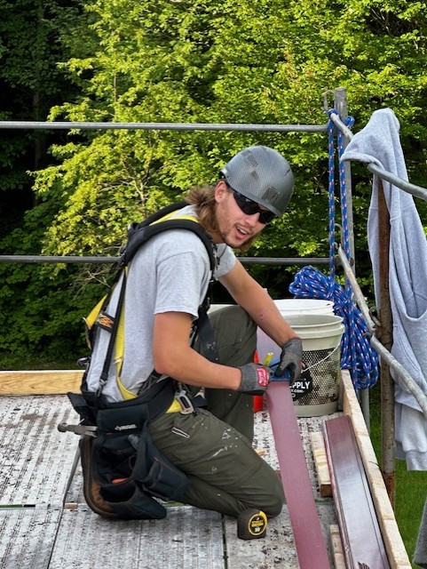 Worker wears dark sunglasses and hardhat kneeling on scaffolding as he marks a plank of wood with a ruler and pencil and looks at camera.