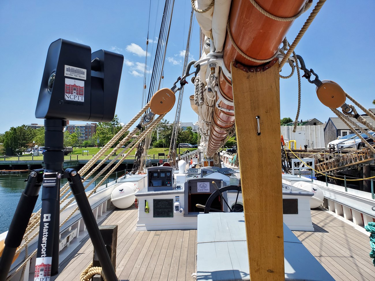 Boxy camera with a domed lens sits on a tripod on the deck of a schooner, just below a boom, facing the wheel and the stern.