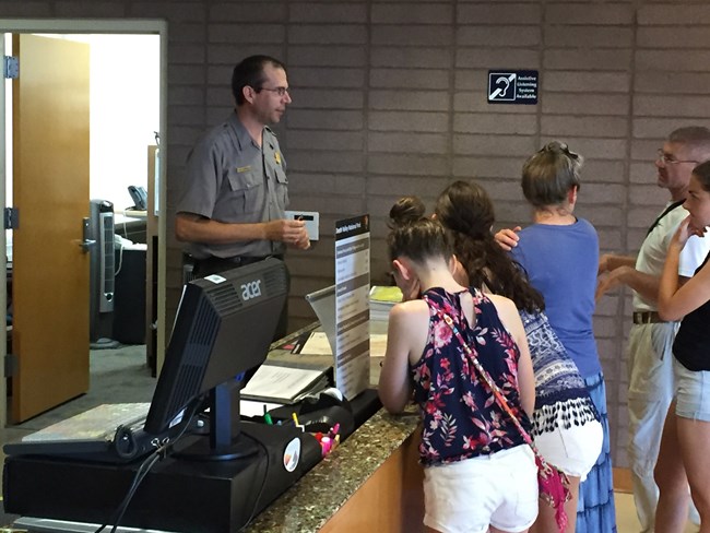 Mike Reynolds wears a ranger uniform and speaks from behind a visitor center desk with park visitors in front of the desk.