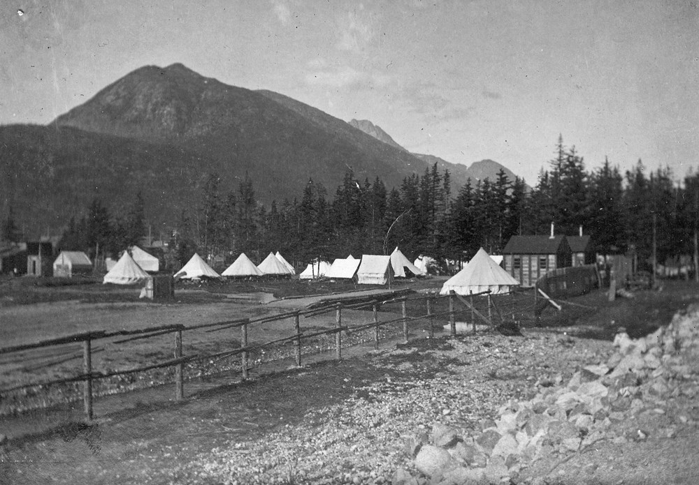 Black and white photo of military camp in Skagway, Alaska. Camp consists of several peaked white canvas tens and a few wooden structures.