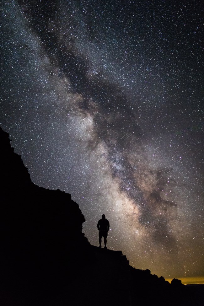 The dark silhouette of a person on a rocky slope, with a very bright night sky and the milky way behind them.