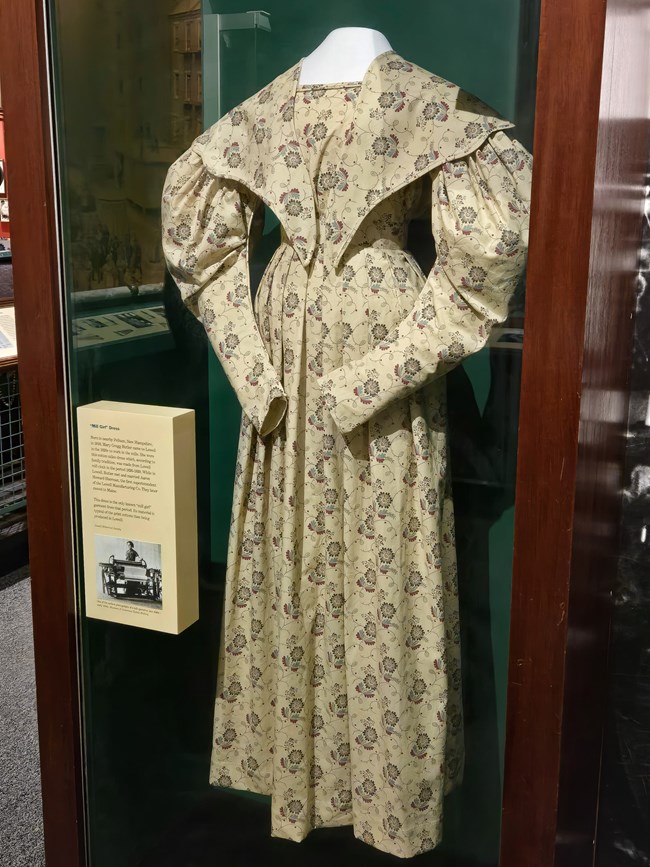 Exhibit case holds a dress made of a yellow fabric with a floral design. The dress features puffy sleeves and a "v" shaped collar