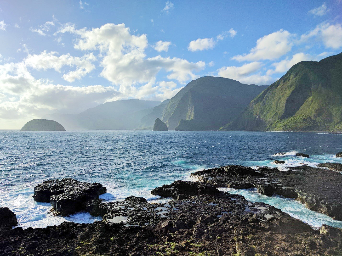 Looking from a rocky, tide-pool-dotted shore across sparkling blue-green water at small islands and towering green sea cliffs, the furthest of which are cloaked in clouds.