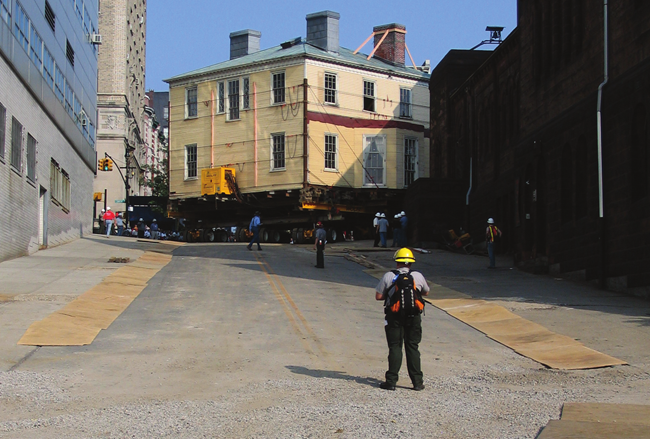 A man stands downhill from Hamilton Grange, which is propped up and moving on hydraulic lifts on the street.