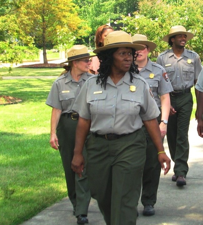 Robin White in her NPS uniform leads a group of uniformed rangers down a sidewalk.