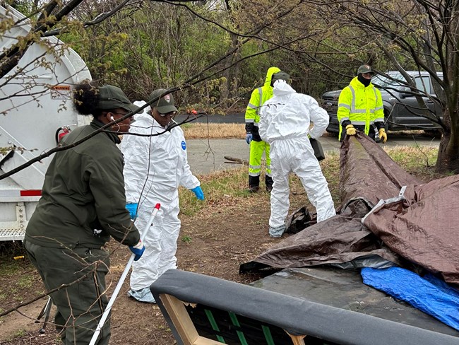 Several maintenance workers wearing protective equipment remove tarps and other objects from an unauthorized encampment.